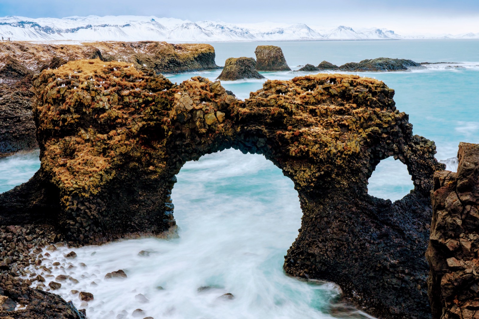Gatklettur, the basalt sea arch near Arnarstapi, with surf running through it and snow-covered peaks behind