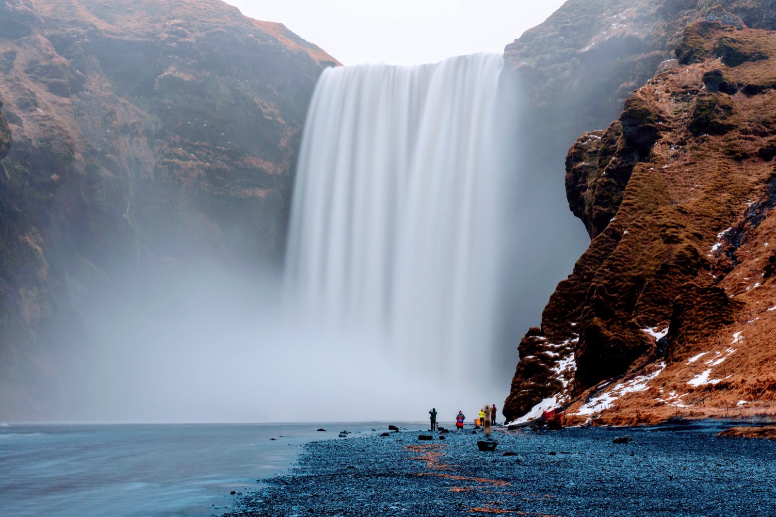 Skógafoss in flat winter light, with photographers on the black gravel at its base for scale