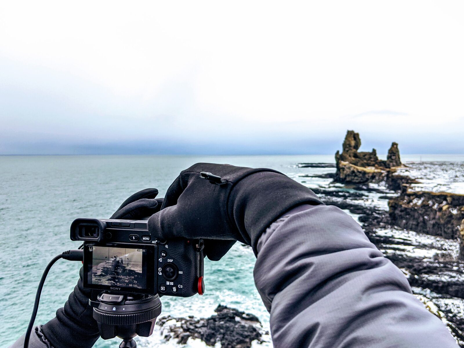 Photographer's gloved hands holding a camera aimed at Lóndrangar